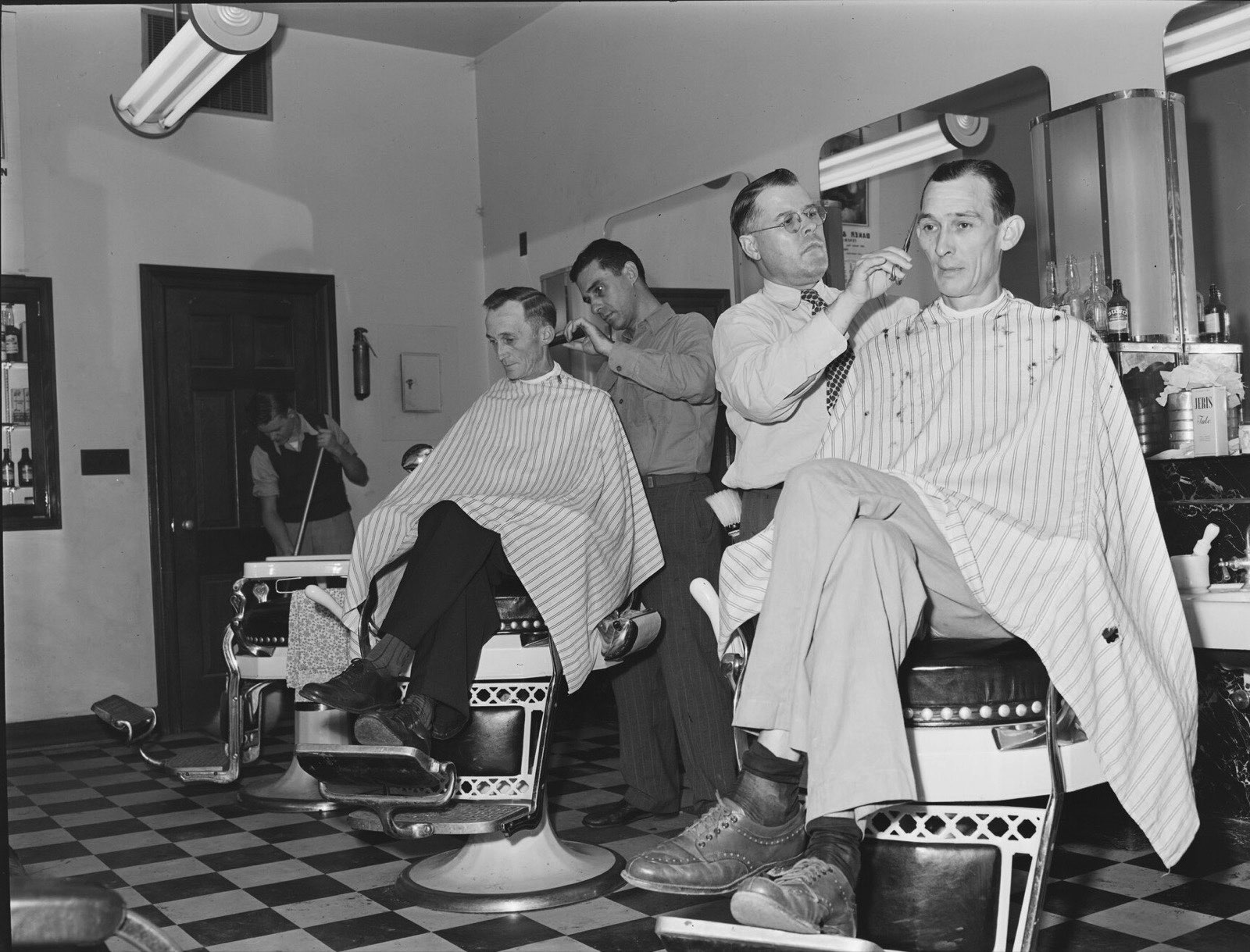 Two barbers cutting the hair of two seated men in striped capes on a checkered floor