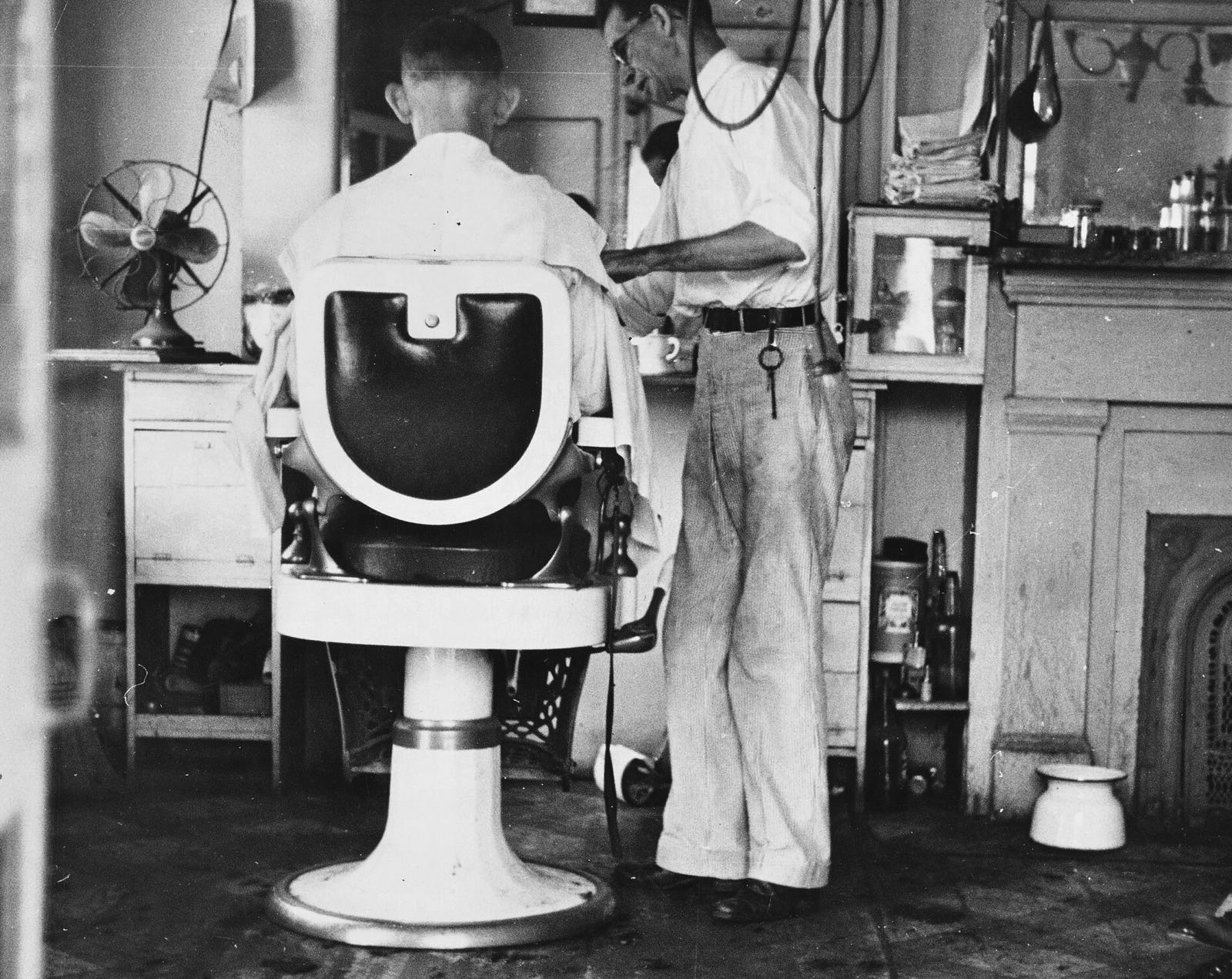 A white enamel barber chair seen from behind, a barber in shirtsleeves at work beside it