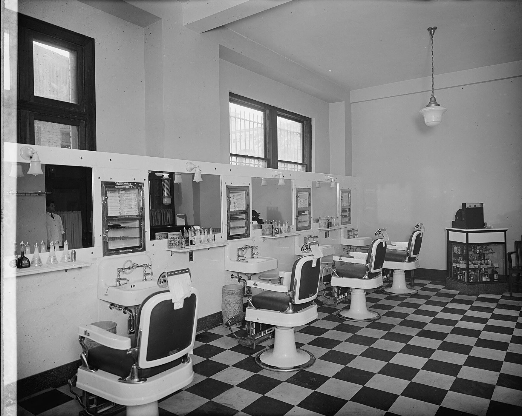 An empty barbershop with a row of white enamel chairs, mirrors and basins along one wall and a black-and-white checkered floor