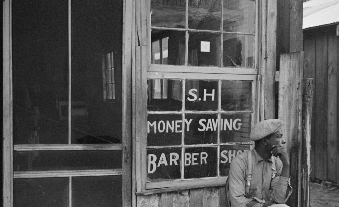 An old barbershop window with hand-painted lettering and a man resting on the step outside