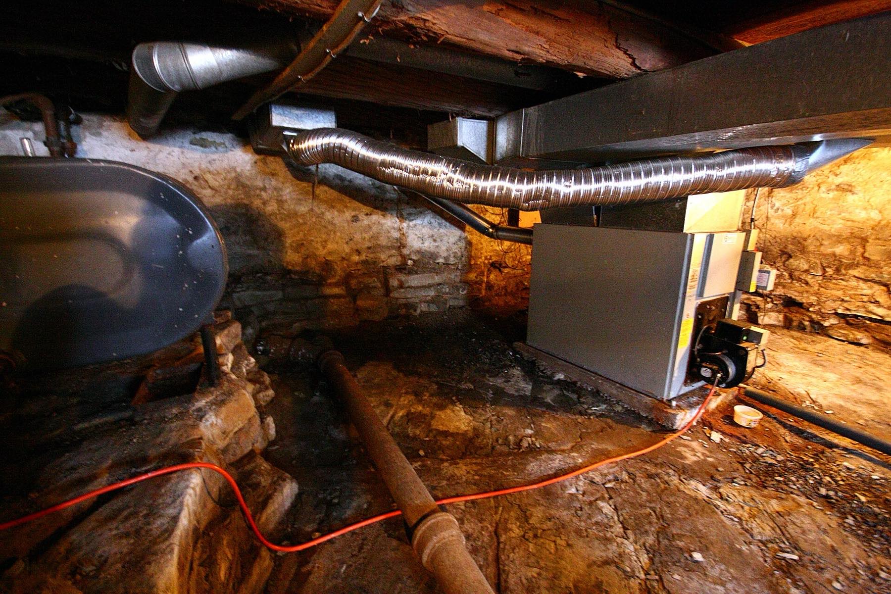 A low stone basement with a furnace, a flexible duct along the joists and an old drain pipe across the floor